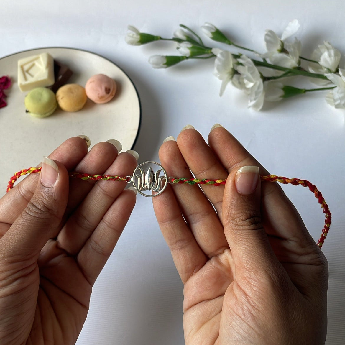 Two hands holding a rakhi bracelet with a decorative lotus on a white background with small flowers and a plate of cookies.