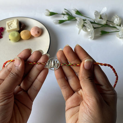 Two hands holding a rakhi bracelet with a decorative lotus on a white background with small flowers and a plate of cookies.